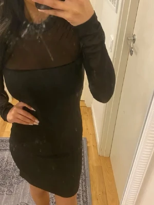 Svart meshklänning med lång ärm - Snygg svart kortklänning med mesh upptill och långa ärmar. Klänningen har en tight passform och en stilren design med transparent parti över bröstet och axlarna. Perfekt för dig som gillar en trendig och elegant look.