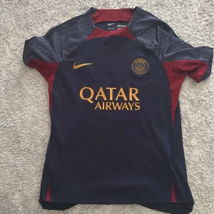 PSG fotbollströja Nike marinblå röd - Paris Saint-Germain matchtröja från Nike i marinblått med röda paneler och gula detaljer. Tryck med Qatar Airways på bröstet, PSG-emblem och Nike-logga. Kortärmad modell i lätt, ventilerande material perfekt för träning eller match.