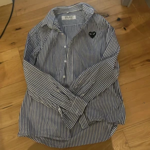 Randig skjorta från Comme des Garçons PLAY - Säljer en blåvit randig skjorta från Comme des Garçons PLAY med klassisk krage och knäppning framtill. Skjortan har det ikoniska svarta hjärta med ögon broderat på bröstet. Perfekt för dig som gillar stilrena och unika detaljer. Kom gärna med prisförslag och eller frågor// Loke