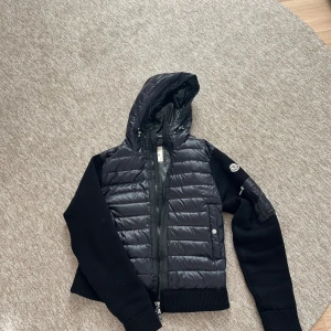 Svart pufferjacka från Moncler - Svart pufferjacka från Moncler med stickade ärmar och huva. Jackan har quiltad front i glansigt material, dragkedja och Moncler-logga på ärmen. Snygg och sportig look med ribbade muddar och praktiska fickor med knappstängning.