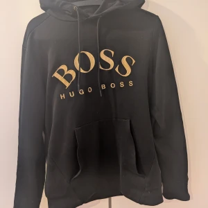 Svart Hugo Boss hoodie  - Svart hoodie från Hugo Boss med stor guldfärgad BOSS-logga på bröstet. Klassisk huva med snörning och stor känguruficka framtill. Väldigt sällsynt men har  Skön passform och perfekt för en snygg streetstyle-look. Skriv för frågor och bilder ⚠️ÄKTA⚠️