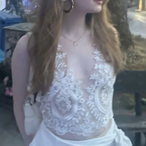  spetsig topp - Säljer en superfin topp i vitt med broderade spetsdetaljer och blommönster. Bortsett från mönstret är den transparent. Dragkedja där bak med knytning runt halsen