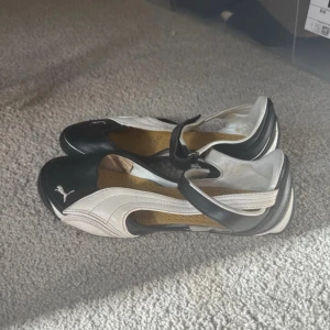 Svartvita ballerinaskor från Puma - Unika svartvita ballerinaskor från Puma med sportig design och klassisk logga på tån. Skorna har rund tå, rem över vristen och är tillverkade i skinn med perforerad innersula för extra komfort. Perfekta för dig som gillar en mix av street och sport.