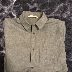 Grå skjorta från HM - Stilren grå skjorta från HM med klassisk krage, bröstficka och svarta knappar. Skjortan har lång ärm och en lätt melerad yta som ger en modern känsla. Perfekt för dig som gillar enkel och clean stil.