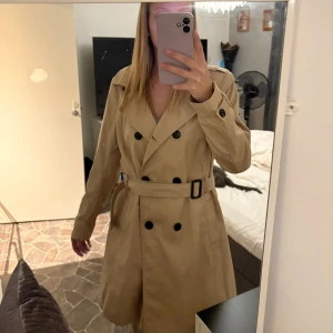 Trenchcoat - Klassisk beige trenchcoat från Stradivarius i storlek XL. Jackan har dubbelknäppning med svarta knappar, bälte i midjan och snygga detaljer vid axlar och ärmslut. Perfekt för lager-på-lager och en clean look.