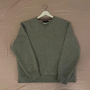 Grå sweatshirt från Tommy Hilfiger - Klassisk grå sweatshirt från Tommy Hilfiger med rund halsringning och diskret broderad logga på bröstet. Tröjan har ribbade muddar vid ärmslut och nederkant, samt en snygg detalj vid halsen. Perfekt för en chill och stilren look.