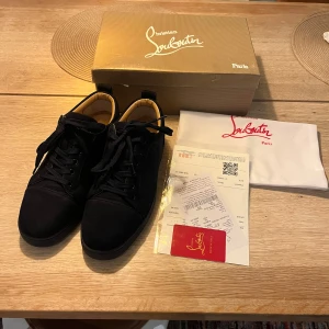 Svarta sneakers från Christian Louboutin - Snygga svarta sneakers från Christian Louboutin i mocka med klassiska röda sulor. Skorna har rund tå, svarta snören och diskret broderad logga på hälen. Insidan är ljusbeige och de levereras med originalkartong och dustbag.