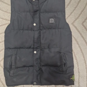 Svart dunväst från Stone Island - Svart dunväst från Stone Island med ikonisk kompasspatch på sidan och logotyp på dragkedjan. Västen har två fickor framtill, tryckknappar och quiltad design. Perfekt för lager-på-lager och streetwear. Snygg, enkel och tidlös look.