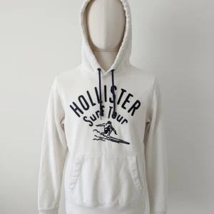 Vit hoodie från Hollister - Vit hoodie från Hollister med svart tryck 'Surf Tour' och surfarmotiv på bröstet. Tröjan har huva med svarta snören och en stor magficka framtill. Perfekt för en avslappnad stil.