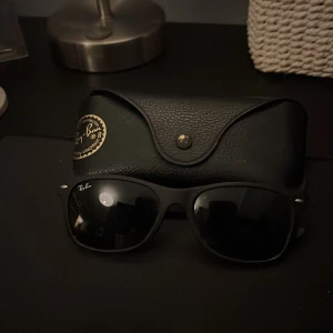 Svarta Ray-Ban Wayfarer solglasögon - Klassiska svarta Ray-Ban Wayfarer solglasögon med ikonisk form och mörka glas. Säljs med original svart fodral i läderlook med Ray-Ban-logga i guld. Perfekt accessoar för en cool och tidlös stil. Nypris 1500