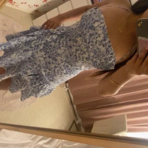 Blommig blåvit kortklänning med volang - Supersöt kort klänning i blått och vitt med blommigt mönster och volangkant. Klänningen har smala axelband och en figurnära passform upptill. Perfekt för varma dagar när du vill känna dig extra fin.