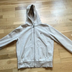 Ljusgrå hoodie från Polo Ralph Lauren - Snygg ljusgrå hoodie från Polo Ralph Lauren med dragkedja framtill, huva med snörning och klassisk broderad logga på bröstet. Tillverkad i mjuk bomullsblandning och har två fickor framtill. Perfekt för en chill och stilren look.