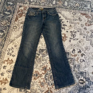 Blå bootcut jeans med pärldetaljer Earl Jean - Snygga blå bootcut jeans från Earl Jean med coola pärl- och strassdetaljer på fickorna. Jeansen har klassisk femficksdesign, kontrastsömmar och är tillverkade i en stretchig bomullsblandning för skön passform.