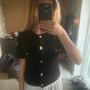 Svart stickad blus med guldknappar - Svart stickad blus med kort ärm och rund hals. Framtill finns två dekorativa fickor med guldiga knappar och hela blusen stängs med matchande guldknappar. Klassisk och stilren modell som är lätt att styla till olika looks. Storlek- 146-152