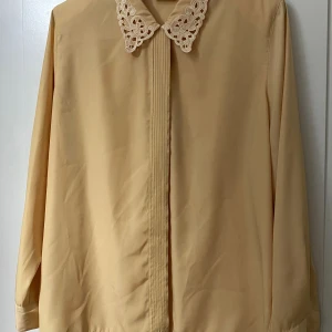 Beige skjorta med spetskrage Elle - Har köpt från secondhand och som binder på bilden är den i bra skick, utan skador men kanske små fläckar som inte syns så mycket. Längden/höjden är 71cm (från axeln till botten), bredden (från ena axeln till den andra) är 43cm och armlängden är 65cm. Meddela mig gärna för mer information!💕💕