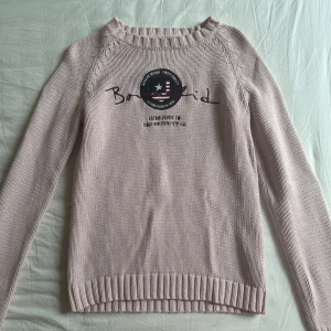 Ljusrosa stickad tröja Bondelid - Ljusrosa stickad tröja från Bondelid med rund halsringning och tryckt logga framtill. Tröjan har ribbade muddar vid ärmslut och nederkant samt diskreta kabelstickade detaljer längs axlarna. Perfekt för lager-på-lager under kyliga dagar.