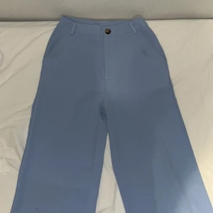 Blå wide kostymbyxor - Snygga ljusblå kostymbyxor med wide fit och hög midja. Byxorna har klassiska fickor fram, bälteshällor och stängs med knapp och dragkedja. Perfekta för dig som gillar en stilren look med lite extra färg.