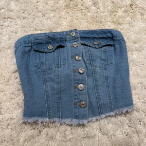 Denim tubtopp med knappar - Säljer en snygg tubtopp i ljusblå denim med silverfärgade knappar framtill, två fejkade bröstfickor och rå fransad nederkant. Baksidan har smock för extra stretch och passform. Perfekt för dig som vill ha en cool och trendig look.