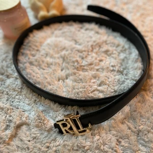Svart skärp från Ralph Lauren - Snyggt svart bälte från Ralph Lauren med stilren design och ett guldigt RL-spänne framtill. Bältet är smalt och gjort i skinn, perfekt för att lyfta vilken outfit som helst med en exklusiv touch. Klassisk och tidlös look med elegant detalj.