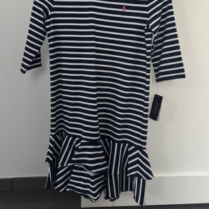 Randig klänning från Polo Ralph Lauren - Snygg randig klänning från Polo Ralph Lauren i marinblått och vitt med rosa logga på bröstet. Modellen har trekvartsärm och volangdetalj nedtill som ger en cool touch. 