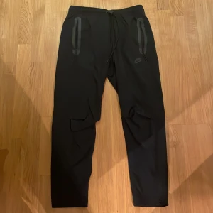 Svarta träningsbyxor från Nike M - Svarta träningsbyxor från Nike med dragsko i midjan och två dragkedjeförsedda fickor framtill. Byxorna har en sportig look, smalare ben och är tillverkade i ett lätt syntetmaterial. Perfekta för träning eller chill. Nike-logga på vänster ben. 