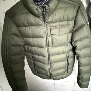 Grön Colmar pufferjacka med dragkedja - Säljer en grön pufferjacka från Colmar med hög krage, dragkedja framtill och fickor med zip. Jackan har ett snyggt Colmar-märke på ärmen och insidan är fodrad i marinblått, beige och vinrött. Perfekt nu till vintern! Hör av dig vid övriga frågor och funderingar!