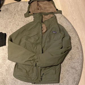 Grön teddyjacka från Patagonia - Säljer en grön teddyjacka från Patagonia med mjukt beige teddyfoder och avtagbar huva. Jackan har dragkedja, två bröstfickor med lock och justerbar nederkant. Perfekt för kalla dagar och har en chill, loose passform.