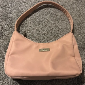 Rosa axelväska från Steve Madden - Snygg ljusrosa axelväska från Steve Madden med stilren design och silverfärgad logodetalj framtill. Väskan har ett huvudfack med dragkedja och en innerficka för småsaker. Materialet är mjuk polyester och remmen är bred och bekväm.