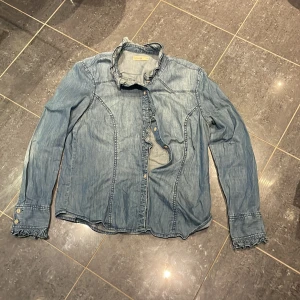 Blå denimskjorta från Denim by Lindex - Snygg blå denimskjorta från Denim by Lindex i storlek 42. Skjortan har långa ärmar med knappar och coola rynkade detaljer vid manschetterna och kragen. Perfekt för dig som gillar avslappnad stil med lite edge.