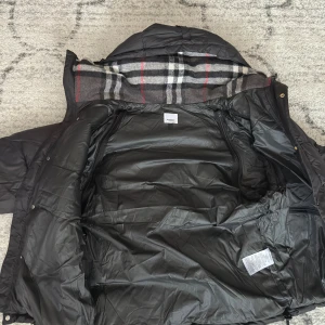 Svart dunjacka från Burberry - Svart dunjacka från Burberry med klassiskt rutigt foder i huvan och tydlig logga på ärmen. Jackan har dragkedja, tryckknappar och två fickor framtill. Perfekt för kalla dagar med sin quiltade design och snygga detaljer.