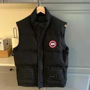 Svart dunväst från Canada Goose - Svart dunväst från Canada Goose med hög krage och klassisk logopatch på bröstet. Västen har två stora fickor framtill med lock och knäppning med både dragkedja och tryckknappar. Perfekt för kalla dagar och har en clean, stilren look.