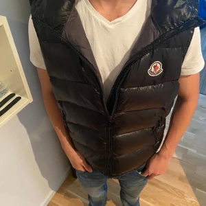 Svart dunväst från Moncler - Snygg svart dunväst från Moncler med glansig finish och klassisk logotyp på bröstet. Västen har hög krage, två sidofickor med dragkedja och quiltad design. Perfekt att slänga över en hoodie eller t-shirt för en cool look.