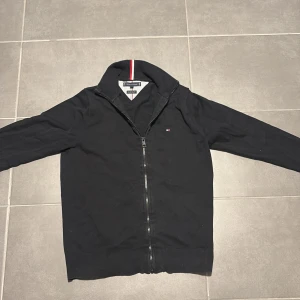 Svart kofta från Tommy Hilfiger - Svart kofta från Tommy Hilfiger med hel dragkedja och långärmad design. Små Hilfiger-loggan på bröstet och snygg krage med röd, vit och blå rand på insidan. Tillverkad i mjuk bomull som är skön att ha på sig. Perfekt för dig som gillar stilrena och klassiska plagg.