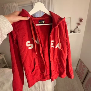Röd hoodie från Svea med dragkedja - Röd hoodie från Svea med vit logga på bröstet, snörning i huvan och praktiska fickor framtill. Tröjan har dragkedja och är långärmad! Materialet är mjuk bomull och passformen är normal. ❤️