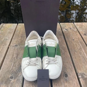 Valentino Garavani Open sneakers vit/grön - Snygga Valentino Garavani Open sneakers i vitt skinn med bred grön läderdetalj över sidan. Klassisk rund tå, platt sula och snörning framtill. Ikoniska nitar på hälen ger extra edge. Perfekt för dig som gillar exklusiva streetwear-vibbar.