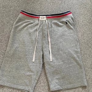 Grå shorts från Polo Ralph Lauren - Snygga grå shorts från Polo Ralph Lauren med klassisk logga broderad på benet. Midjan har röd och marinblå rand samt snörning för perfekt passform. Mjukt bomullsmaterial som är skönt mot huden, perfekt för chill dagar eller träning.