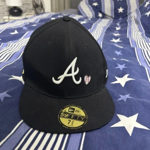 Svart New Era 59FIFTY keps 7 3/4 - Säljer en svart New Era 59FIFTY keps med broderad vit 'A' och ett litet rosa hjärta framtill. Klassisk flat peak och MLB Genuine Merchandise-märke på insidan. Perfekt för dig som gillar streetwear och sportig stil.