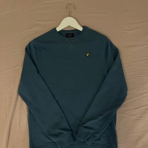 Blå sweatshirt från Lyle & Scott - Snygg blå sweatshirt från Lyle & Scott med klassisk rund hals och broderad gul logga på bröstet. Tröjan har långa ärmar och ribbade muddar vid ärmslut och nederkant. Perfekt för en chill och stilren look.