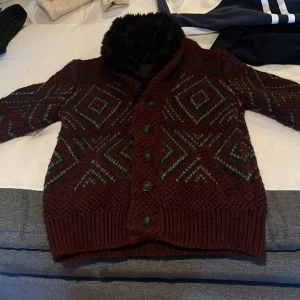 Vinröd stickad cardigan jacka med mönster - Mysig vinröd cardigan jacka med stickat rutmönster i grått och svart. Jackan har stora knappar framtill, två fickor och en fluffig svart fuskpälskrage. Perfekt för kalla dagar och ger en chill vibe till din outfit.