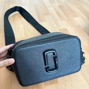 Svart axelväska från Marc Jacobs - Snygg svart axelväska från Marc Jacobs med bred justerbar axelrem och präglad logga framtill. Väskan är tillverkad i strukturerat skinn och har dragkedja upptill samt ett praktiskt ytterfack på baksidan. Perfekt för att bära det viktigaste med stil.