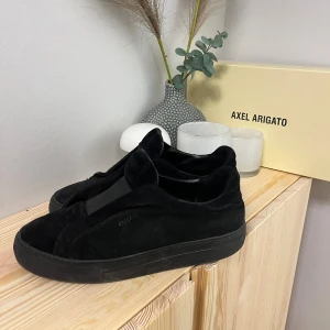 Svarta sneakers från Axel Arigato - Säljer ett par svarta sneakers från Axel Arigato i storlek 41. Skorna har en stilren design med rund tå, platt sula och är tillverkade i mocka med detaljer i skinn. De har elastiska paneler istället för klassisk snörning för en clean look. lite snöre har gått upp inne i skon, kom privat för frågor🔥