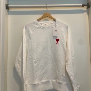 Vit sweatshirt från Ami med rött hjärta - Vit sweatshirt från Ami Alexandre Mattiussi med klassisk rund hals och lång ärm. På bröstet finns ett broderat rött hjärta med bokstaven A. Tröjan är i mjuk bomull och har en clean, stilren look som passar perfekt till jeans eller dressade byxor.