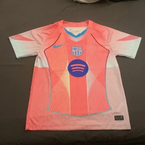 Barcelona rosa/blå Nike fotbollströja - Säljer en Barcelona fotbollströja från Nike i en cool rosa och ljusblå färg med grafiskt mönster. Tröjan har korta ärmar, V-ringning och tryck med Spotify-logga och klubbmärke på bröstet. Materialet är lätt och andas, perfekt för match eller träning.