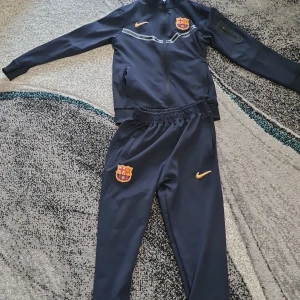 Nike FC Barcelona träningsbyxor 154 - Mörkblå träningsbyxor från Nike med FC Barcelona-logga och gul Nike-swoosh på benet. Byxorna har resår i midjan, smal passform och muddar vid benslut. Perfekta för fotbollsträning eller chill. Materialet är syntet och känns lätt och smidigt.