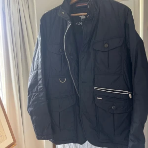 Mörkblå quiltad jacka från Snoot - Mörkblå/navy quiltad jacka från Snoot med flera fickor framtill, dragkedja och knappar. Jackan har hög krage, detaljer i silver och är tillverkad i Italien. Innerväst som inte finns med på bild ingår även i köpet. Kan skicka bild på begäran. Förvånadsvärt varm jacka så funkar på vintern också, annars perfekt för hösten.