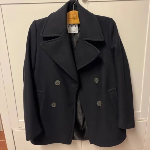 Svart kappa från Zara - Stilren svart kappa från Zara med dubbelknäppning och bred krage. Jackan har två fickor framtill och är fodrad på insidan. Klassisk modell som passar perfekt till höst och vinter. Materialet är ullblandning för en varm känsla. Passar perfekt till både hösten och vintern. Jackan är aldrig använd men finns ingen prislapp kvar. 