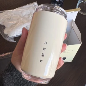 Smeg Wall Mug - Elegant Smeg Wall Mug i krämfärgad design. Perfekt för varma och kalla drycker, med lock för spillfri användning. Ser ut att vara i nyskick, levereras med originalförpackning. Stilren och praktisk för både hem och resa. 240ml