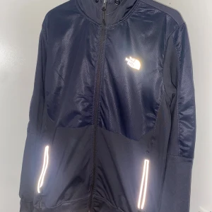 North Face vindjacka - Mörk blå vindjacka från The North Face med reflekterande detaljer vid fickorna och logga på bröstet. Jackan har dragkedja hela vägen, hög krage och justerbar huva. Tillverkad i polyester och har en sportig, clean look som passar perfekt till streetwear.