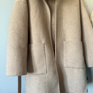 Beige kappa med päls och bälte - Säljer en beige kappa med stor fluffig päls runt kragen och praktiskt bälte i midjan. Jackan har två stora fickor framtill och är tillverkad i mjukt ullmaterial. Perfekt för dig som vill ha en stilren och mysig look under kyliga dagar.