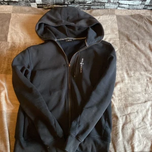 Svart hoodie från Sail Racing - Svart hoodie från Sail Racing med dragkedja framtill och en bröstficka med logga. Tröjan har huva, långa ärmar och är fodrad med mjuk fleece på insidan. Perfekt för kyliga dagar och snygg till jeans eller joggers.